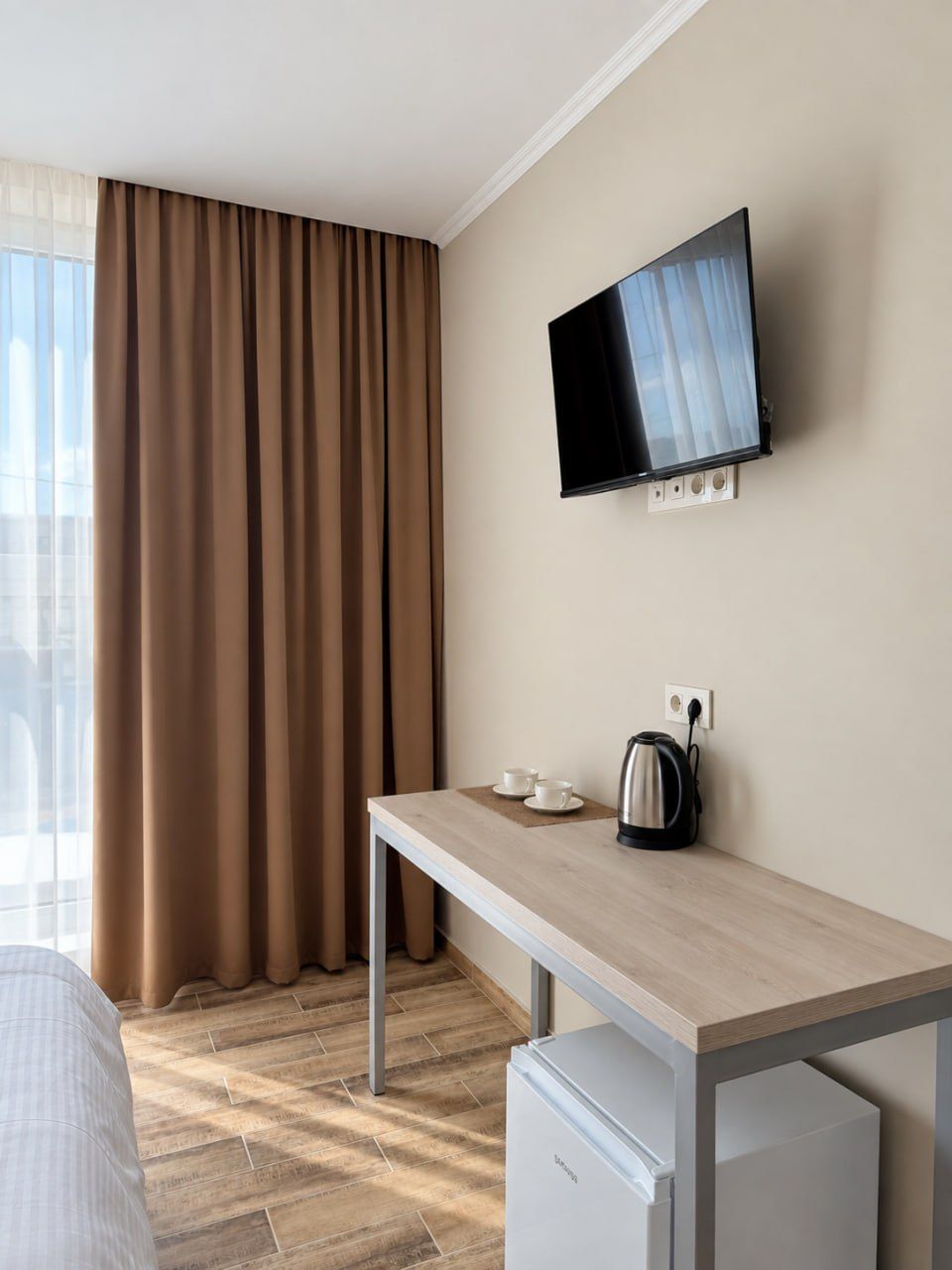 Birou cu fierbător și două cești, televizor pe perete, draperii maro și mini-frigider sub birou